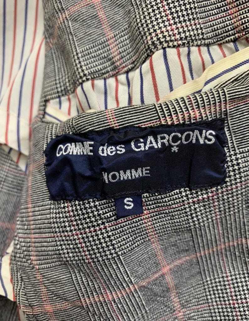 Op de afbeelding: Detail van een blazerinterieur met een zwart, wit en rood ruitpatroon. De voering heeft rode en blauwe strepen op een witte achtergrond. Een marineblauw label leest "COMME des GARCONS HOMME" met een maat "S" tag.