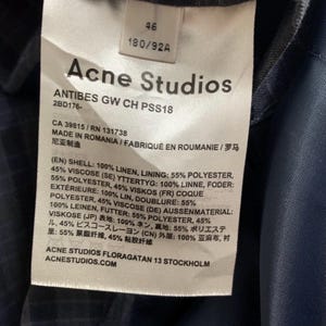 May include: A white tag with the Acne Studios logo and product information. The tag includes details about the garment's composition, such as 100% linen and 55% polyester. The tag also includes the size 46 and the measurements 180/92A.