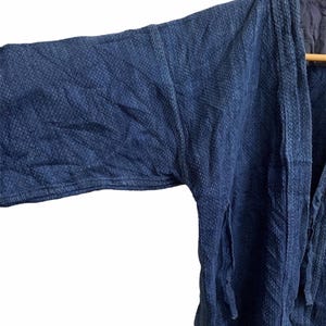 May include: Close-up of a dark blue, textured kimono-style jacket. The jacket has long sleeves and a relaxed fit. The fabric appears to be a woven material with a subtle pattern. The jacket is hanging on a wooden hanger.