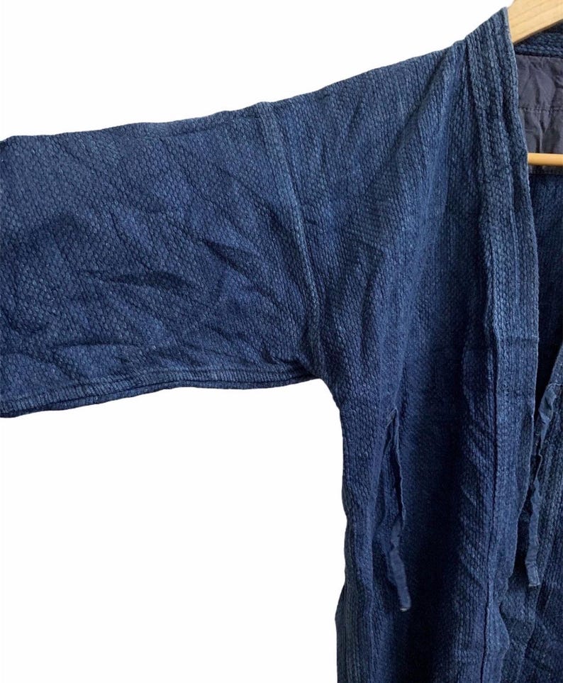 May include: Close-up of a dark blue, textured kimono-style jacket. The jacket has long sleeves and a relaxed fit. The fabric appears to be a woven material with a subtle pattern. The jacket is hanging on a wooden hanger.