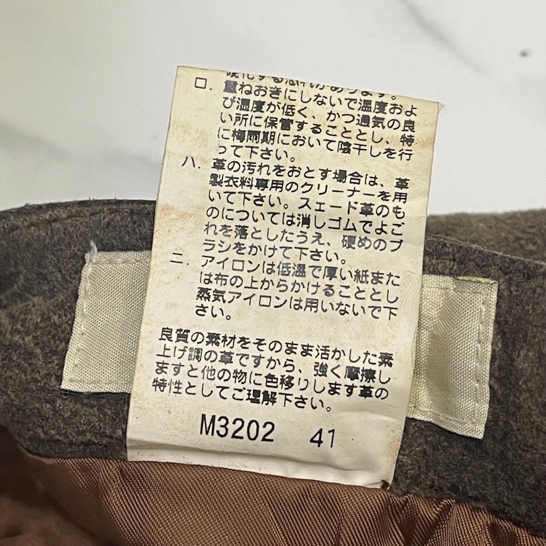 Pode incluir: Close-up de um tecido castanho com uma etiqueta branca anexada. A etiqueta tem texto japon&ecirc;s e os n&uacute;meros "M3202" e "41" impressos. O tecido parece fazer parte de uma pe&ccedil;a de vestu&aacute;rio.