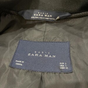 Puede incluir: Primer plano del forro de una chaqueta verde oscuro con dos etiquetas azul marino. Las etiquetas dicen "BASIC ZARA MAN" e incluyen informaci&oacute;n de tallas: EUR L, USA L y MEX 40. La chaqueta est&aacute; hecha en China.