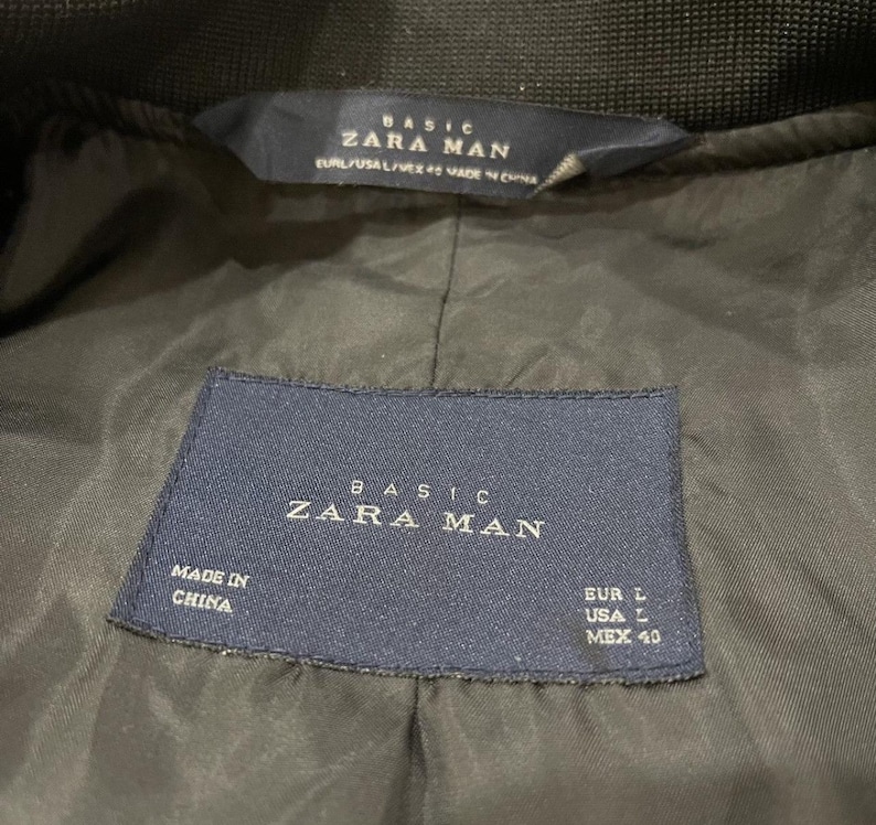 Puede incluir: Primer plano del forro de una chaqueta verde oscuro con dos etiquetas azul marino. Las etiquetas dicen "BASIC ZARA MAN" e incluyen informaci&oacute;n de tallas: EUR L, USA L y MEX 40. La chaqueta est&aacute; hecha en China.
