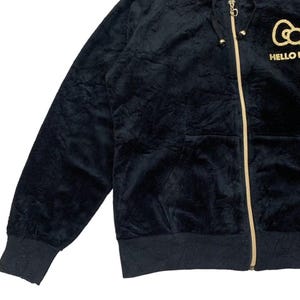 Puede incluir: Sudadera con capucha negra con cremallera dorada y el texto "HELLO KITTY" en dorado. La sudadera tiene una textura suave y aterciopelada y un bolsillo delantero. El logotipo de Hello Kitty est&aacute; bordado en el pecho.