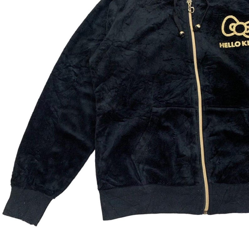 Puede incluir: Sudadera con capucha negra con cremallera dorada y el texto "HELLO KITTY" en dorado. La sudadera tiene una textura suave y aterciopelada y un bolsillo delantero. El logotipo de Hello Kitty est&aacute; bordado en el pecho.