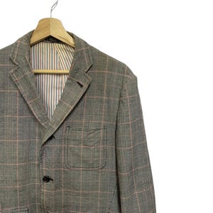 Op de afbeelding: Een grijze geruite blazer met een roze rasterpatroon, hangend aan een houten hanger. De blazer heeft een gekerfde revers, een borstzak en drie zwarte knopen. De binnenvoering heeft verticale strepen in blauw, rood en wit.