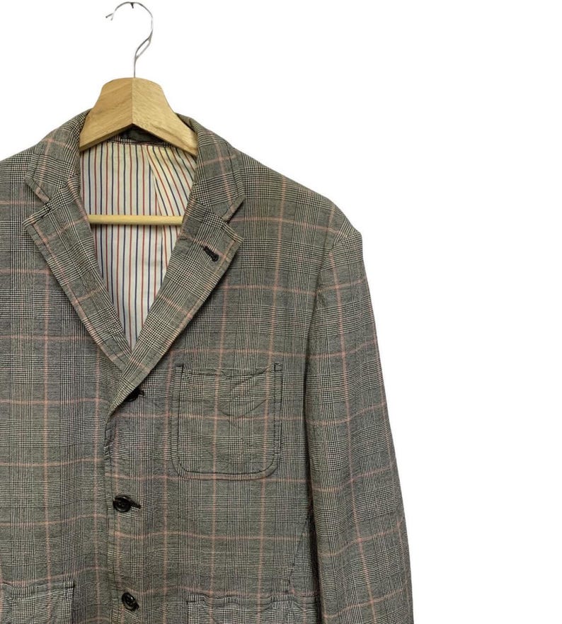 Op de afbeelding: Een grijze geruite blazer met een roze rasterpatroon, hangend aan een houten hanger. De blazer heeft een gekerfde revers, een borstzak en drie zwarte knopen. De binnenvoering heeft verticale strepen in blauw, rood en wit.