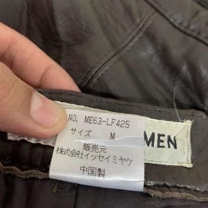 May include: Close-up of a brown leather garment with a tag. The tag displays the product number ME63-LF425, size M, and the brand name. The tag also includes Japanese text and the words "Made in China."