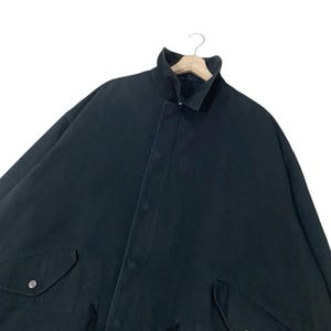 Puede incluir: Chaqueta negra con cuello alto y cierre de botones. La chaqueta tiene dos bolsillos delanteros con cierres de bot&oacute;n plateados. La prenda est&aacute; colgada de una percha de madera sobre un fondo blanco.