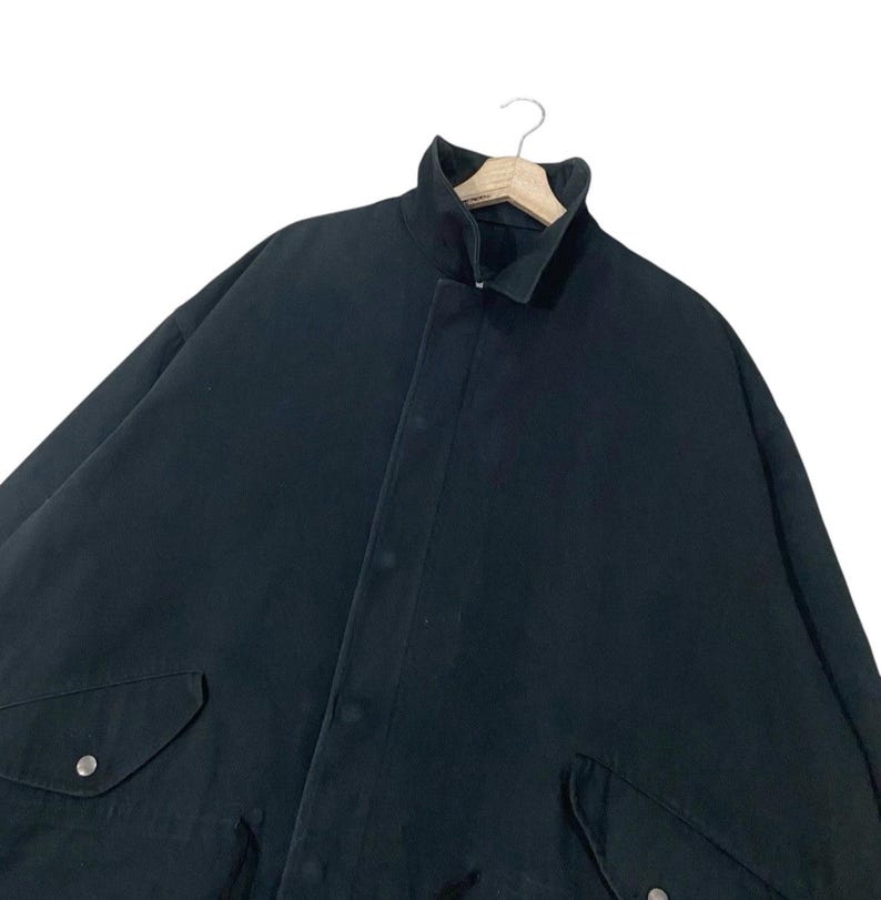 Puede incluir: Chaqueta negra con cuello alto y cierre de botones. La chaqueta tiene dos bolsillos delanteros con cierres de bot&oacute;n plateados. La prenda est&aacute; colgada de una percha de madera sobre un fondo blanco.