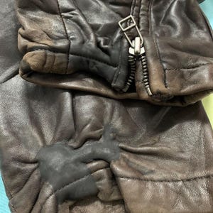 May include: Close-up of a distressed brown leather jacket. The jacket features a silver zipper and a worn appearance, with visible creases and color variations. The leather shows signs of wear and tear.