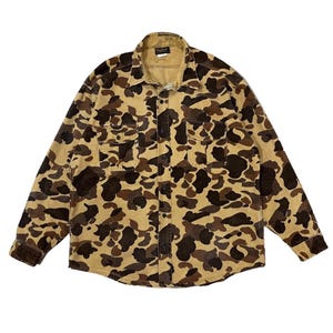 May include: A long-sleeved button-down shirt in a camouflage pattern. The shirt features a classic collar, two chest pockets, and a button-up front. The camouflage design incorporates shades of brown, tan, and black.
