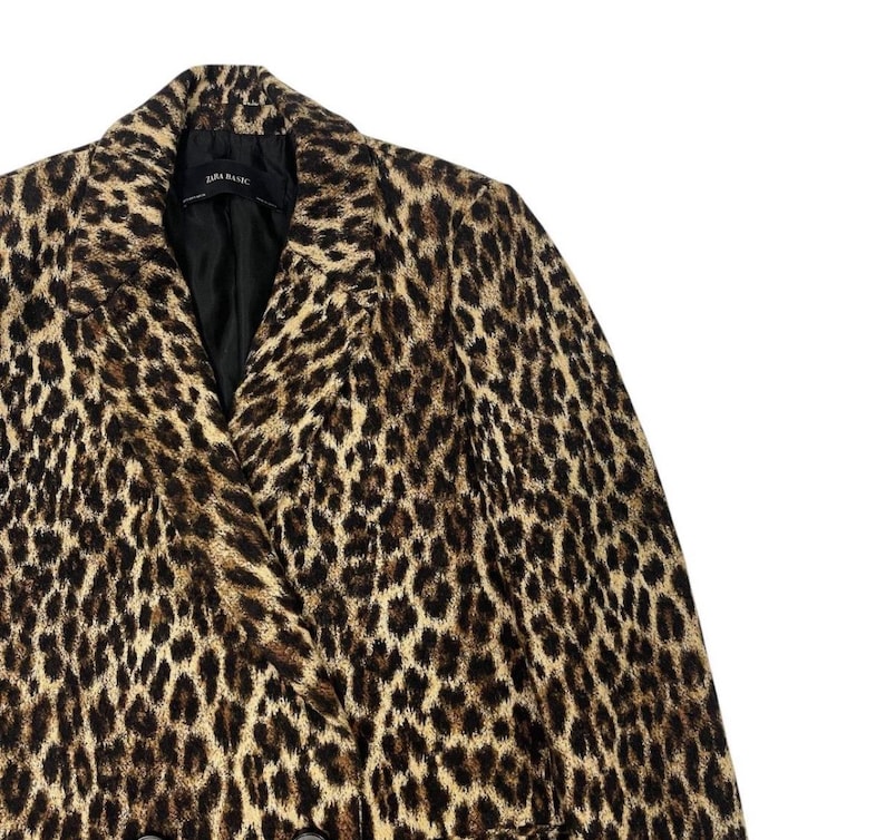 May include: A leopard print blazer with a black lining. The jacket features a classic notched collar and a double-breasted design. The print is a mix of tan, brown, and black spots. The label reads "ZARA BASIC".