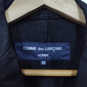 May include: Close-up of a black jacket with a navy blue label. The label reads "COMME des GAR&Ccedil;ONS HOMME" in white text. Below the text is the size "SS". The jacket is hanging on a wooden hanger.