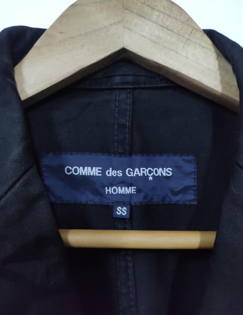 May include: Close-up of a black jacket with a navy blue label. The label reads "COMME des GAR&Ccedil;ONS HOMME" in white text. Below the text is the size "SS". The jacket is hanging on a wooden hanger.