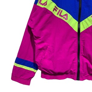 May include: A vibrant, retro-style jacket in shades of fuchsia, blue, and neon yellow. The jacket features a full zipper and the brand name "FILA" in a bold, pink font. The design includes color-blocked panels and stripes, creating a sporty aesthetic.