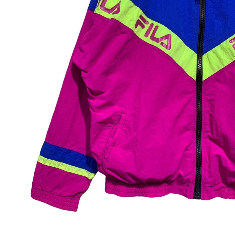 May include: A vibrant, retro-style jacket in shades of fuchsia, blue, and neon yellow. The jacket features a full zipper and the brand name "FILA" in a bold, pink font. The design includes color-blocked panels and stripes, creating a sporty aesthetic.