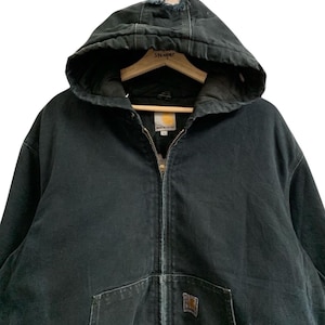 May include: Black hooded Carhartt jacket with a zipper closure and front pockets. The jacket has a worn appearance, with distressed areas on the hood. A small Carhartt logo patch is visible on the front pocket. The jacket is hanging on a wooden hanger.