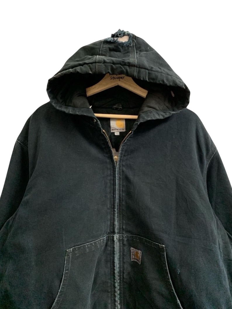 May include: Black hooded Carhartt jacket with a zipper closure and front pockets. The jacket has a worn appearance, with distressed areas on the hood. A small Carhartt logo patch is visible on the front pocket. The jacket is hanging on a wooden hanger.