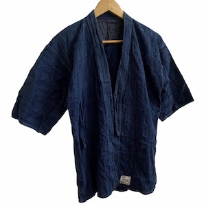 May include: A dark blue, open-front jacket with three-quarter sleeves, hanging on a wooden hanger. The jacket has a textured appearance and a small label near the bottom. The garment is likely made of a durable fabric.