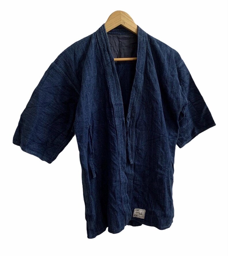 May include: A dark blue, open-front jacket with three-quarter sleeves, hanging on a wooden hanger. The jacket has a textured appearance and a small label near the bottom. The garment is likely made of a durable fabric.