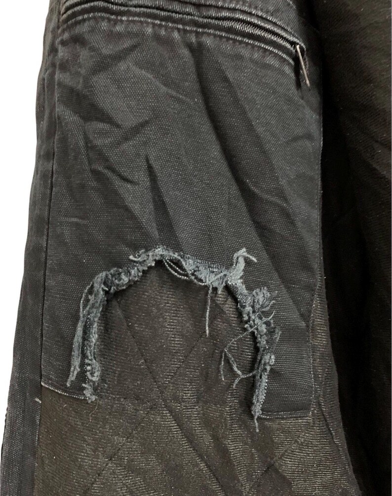 May include: Close-up of a black jacket interior with a torn pocket. The pocket is quilted with a diamond pattern. A zipper is visible at the top. The jacket appears to be made of a durable fabric.