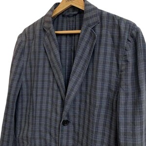 May include: A blue and gray plaid blazer with a single black button closure. The jacket is hanging on a wooden hanger. The blazer has a classic notched lapel and a relaxed fit.