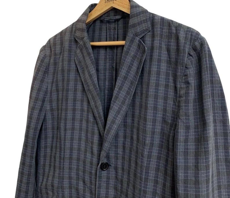 May include: A blue and gray plaid blazer with a single black button closure. The jacket is hanging on a wooden hanger. The blazer has a classic notched lapel and a relaxed fit.