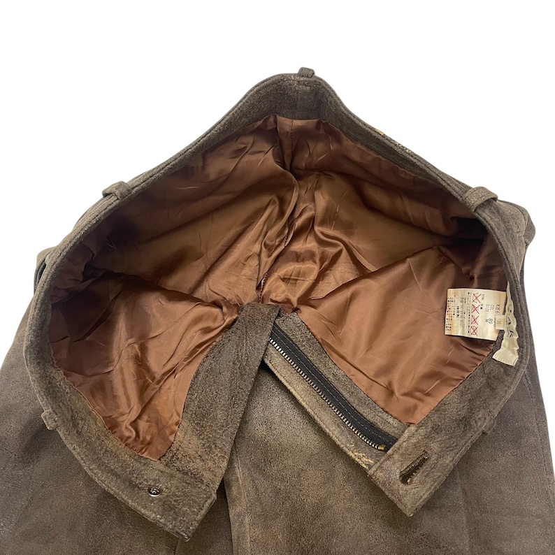 Pode incluir: Um par de cal&ccedil;as de camur&ccedil;a castanhas, visto por dentro. As cal&ccedil;as t&ecirc;m forro de cetim castanho e fecho de correr. A cintura tem presilhas e fecho de bot&atilde;o. Uma etiqueta com texto &eacute; vis&iacute;vel.