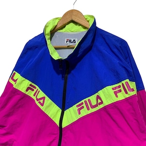 May include: A vintage Fila track jacket in blue, pink, and neon green. The jacket features a full zipper, a stand-up collar, and the Fila logo on the chest and across the chest in a chevron design. The jacket is hanging on a wooden hanger.