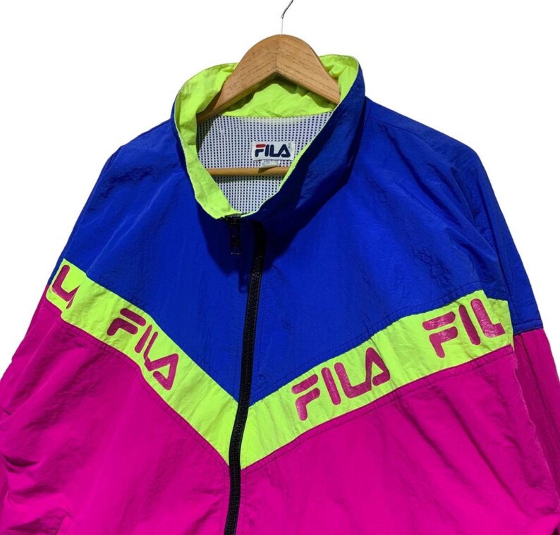 May include: A vintage Fila track jacket in blue, pink, and neon green. The jacket features a full zipper, a stand-up collar, and the Fila logo on the chest and across the chest in a chevron design. The jacket is hanging on a wooden hanger.