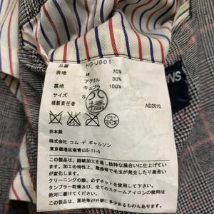 Op de afbeelding: Close-up van een kledinglabel met Japanse tekst en details. Het label bevat percentages van de stofcompositie, maat en wasvoorschriften. De achtergrond toont een geruite stof.