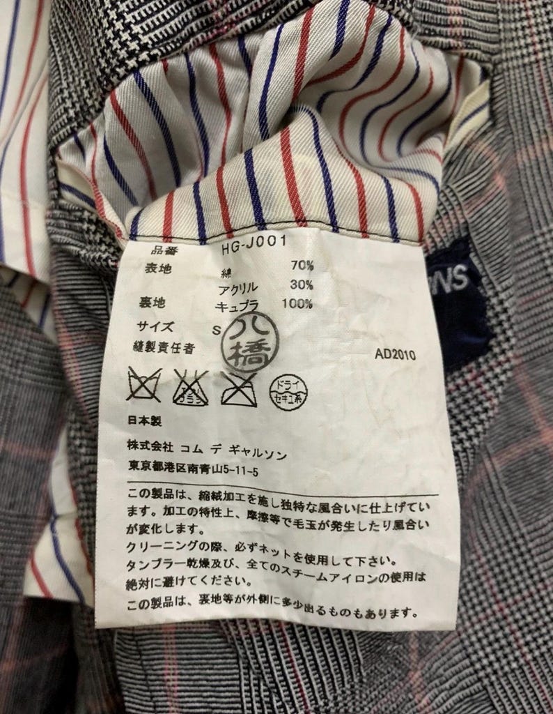 Op de afbeelding: Close-up van een kledinglabel met Japanse tekst en details. Het label bevat percentages van de stofcompositie, maat en wasvoorschriften. De achtergrond toont een geruite stof.