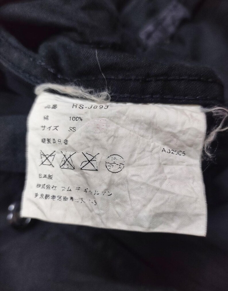 May include: Close-up of a white clothing tag with Japanese text and symbols, including "HS-J093", "100%", and "SS". The tag is attached to a dark fabric, with the code "AJ2005" visible.