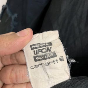 May include: A close-up of a Carhartt clothing tag. The tag is white with black text that reads "Produced By UFCW Union Labor Carhartt". The tag is attached to a black garment.