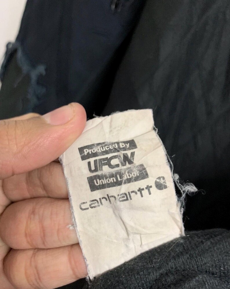 May include: A close-up of a Carhartt clothing tag. The tag is white with black text that reads "Produced By UFCW Union Labor Carhartt". The tag is attached to a black garment.