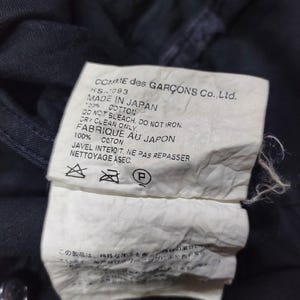 May include: Close-up of a dark navy garment tag. The tag reads "COMME des GAR&Ccedil;ONS Co., Ltd. MADE IN JAPAN" with care instructions. The fabric is 100% cotton. The tag is white with black text.