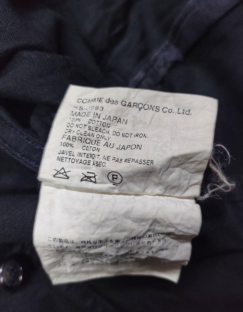 May include: Close-up of a dark navy garment tag. The tag reads "COMME des GAR&Ccedil;ONS Co., Ltd. MADE IN JAPAN" with care instructions. The fabric is 100% cotton. The tag is white with black text.