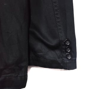 May include: Close-up of a black blazer. The blazer has three black buttons on the sleeve cuff. The fabric appears to be a smooth, solid color. The stitching is visible along the seams.