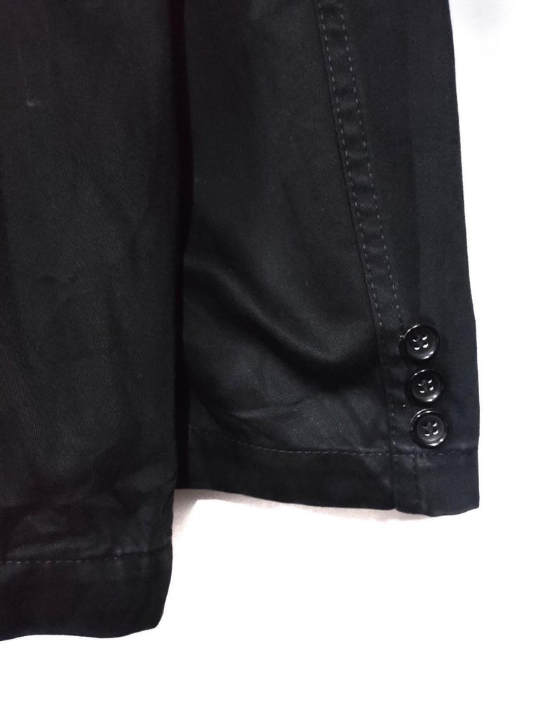 May include: Close-up of a black blazer. The blazer has three black buttons on the sleeve cuff. The fabric appears to be a smooth, solid color. The stitching is visible along the seams.