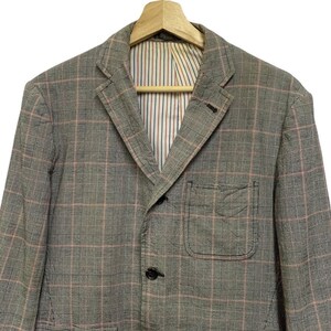 Op de afbeelding: Een grijze blazer met een roze en wit ruitpatroon. De blazer heeft een revers, twee voorzakken en drie knopen. De binnenvoering heeft een gestreept patroon in rood, wit en blauw.