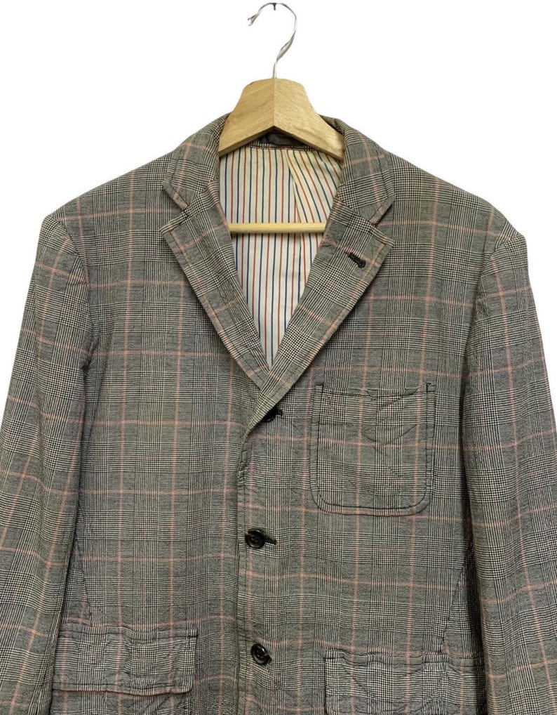 Op de afbeelding: Een grijze blazer met een roze en wit ruitpatroon. De blazer heeft een revers, twee voorzakken en drie knopen. De binnenvoering heeft een gestreept patroon in rood, wit en blauw.