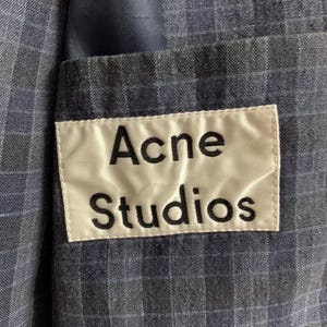 May include: Close-up of a blue plaid blazer with a white label that reads "Acne Studios" in black letters. The blazer has a pocket and a blue lining.