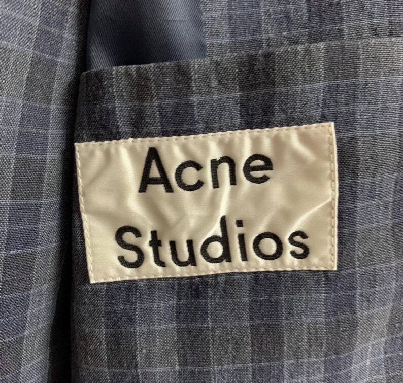 May include: Close-up of a blue plaid blazer with a white label that reads "Acne Studios" in black letters. The blazer has a pocket and a blue lining.