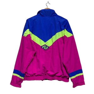 May include: A vintage Fila jacket in a vibrant color block design. The jacket features a combination of bright pink, royal blue, and neon green. The Fila logo is visible on the chest. The jacket has a zip-up front and a high collar.
