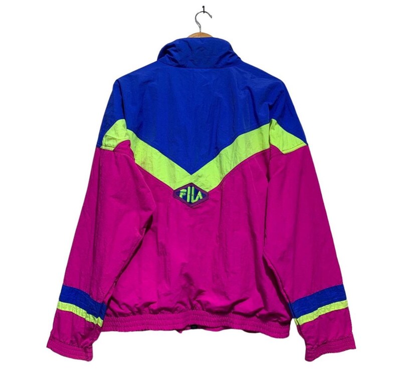 May include: A vintage Fila jacket in a vibrant color block design. The jacket features a combination of bright pink, royal blue, and neon green. The Fila logo is visible on the chest. The jacket has a zip-up front and a high collar.