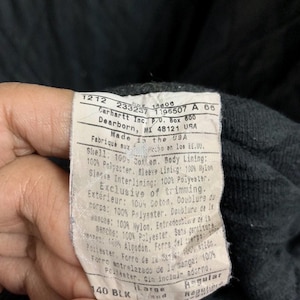 May include: Close-up of a Carhartt label with text. The label is white with black text and includes the company's address in Dearborn, MI. The label also lists the materials used, including cotton, polyester, and nylon. The size is Large.