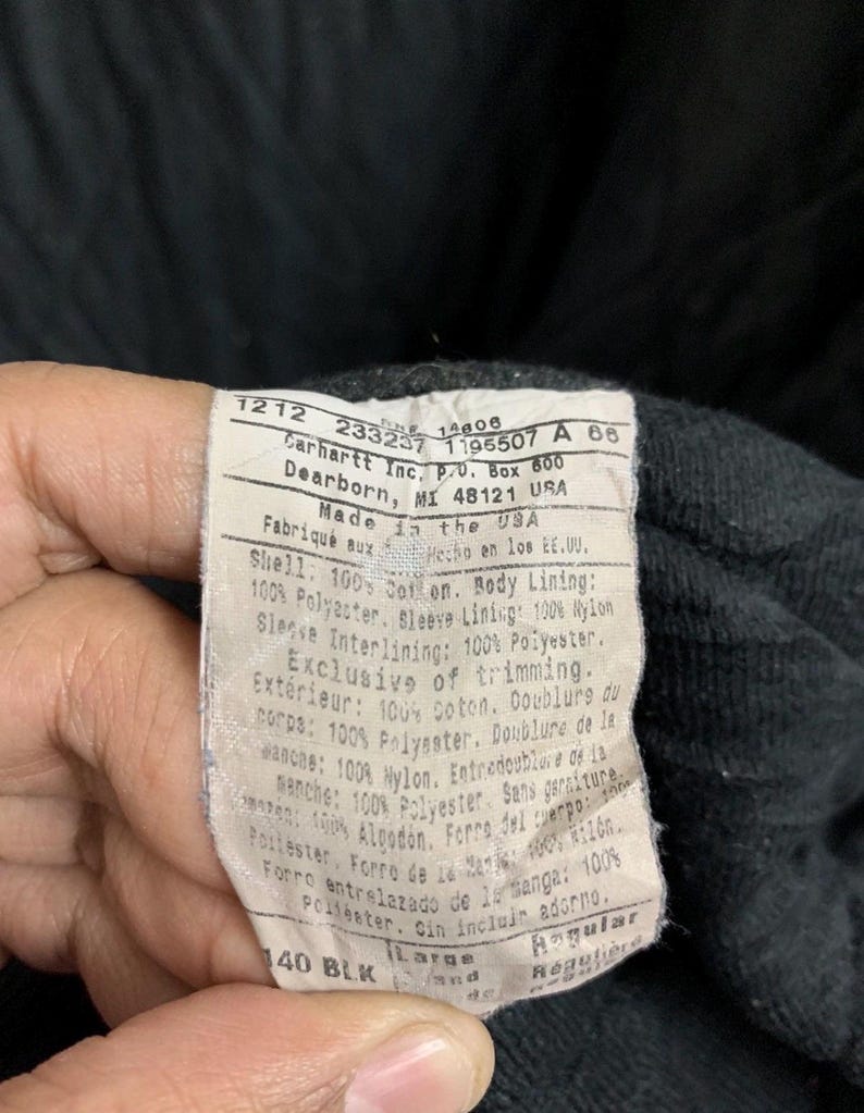 May include: Close-up of a Carhartt label with text. The label is white with black text and includes the company's address in Dearborn, MI. The label also lists the materials used, including cotton, polyester, and nylon. The size is Large.