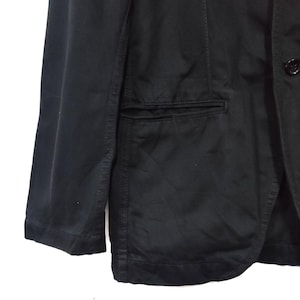 May include: A black blazer with a single-breasted design. The blazer features a notched lapel, a chest pocket, and two buttons. The sleeves are long and the fabric appears to be a cotton blend.