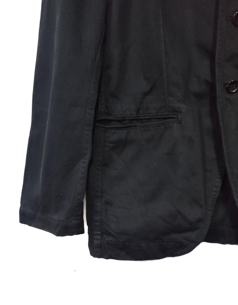 May include: A black blazer with a single-breasted design. The blazer features a notched lapel, a chest pocket, and two buttons. The sleeves are long and the fabric appears to be a cotton blend.